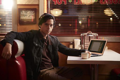 Jughead sitting in a restaurant booth with a laptop open to a blank word document