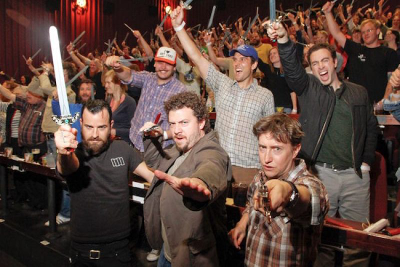 Actors Justin Theroux, Danny McBride, and director David Gordon Green stand in front of a crowd of moviegoers all holding swords at a movie event.