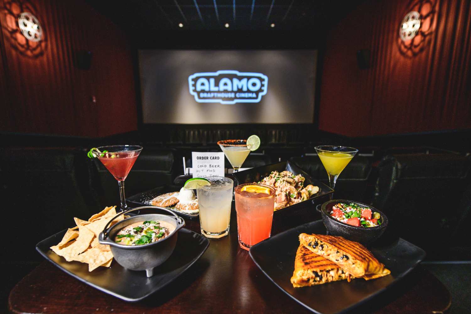 Several plates of food and alcoholic beverages sit on a table in an Alamo Drafthouse theater.