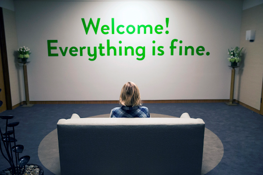 Kristen Bell is seen from the back, sitting on a couch facing a wall with the words 'Welcome! Everything is fine.' printed in green.
