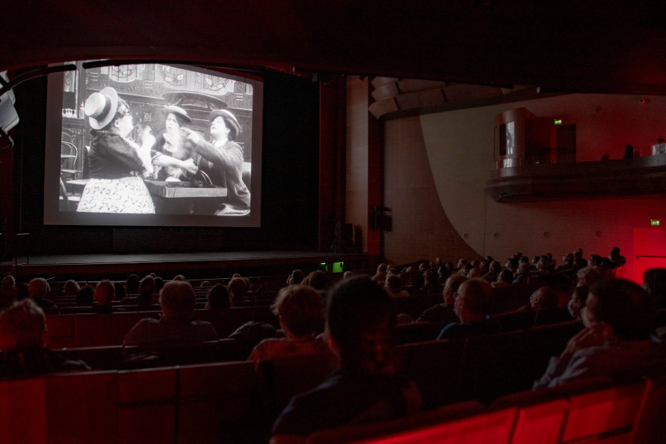audience in a movie theater watches black and white film with three women fighting on screen