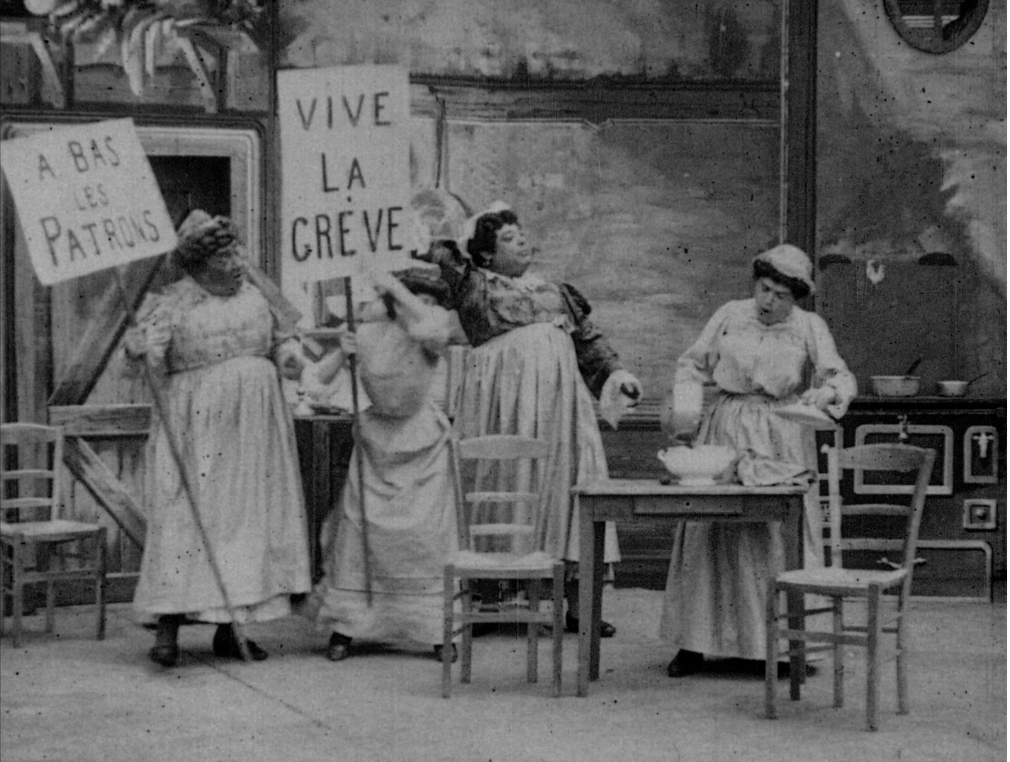 Screencap from a silent film in which four maids are protesting in a kitchen, two of whom are holding signs that say 