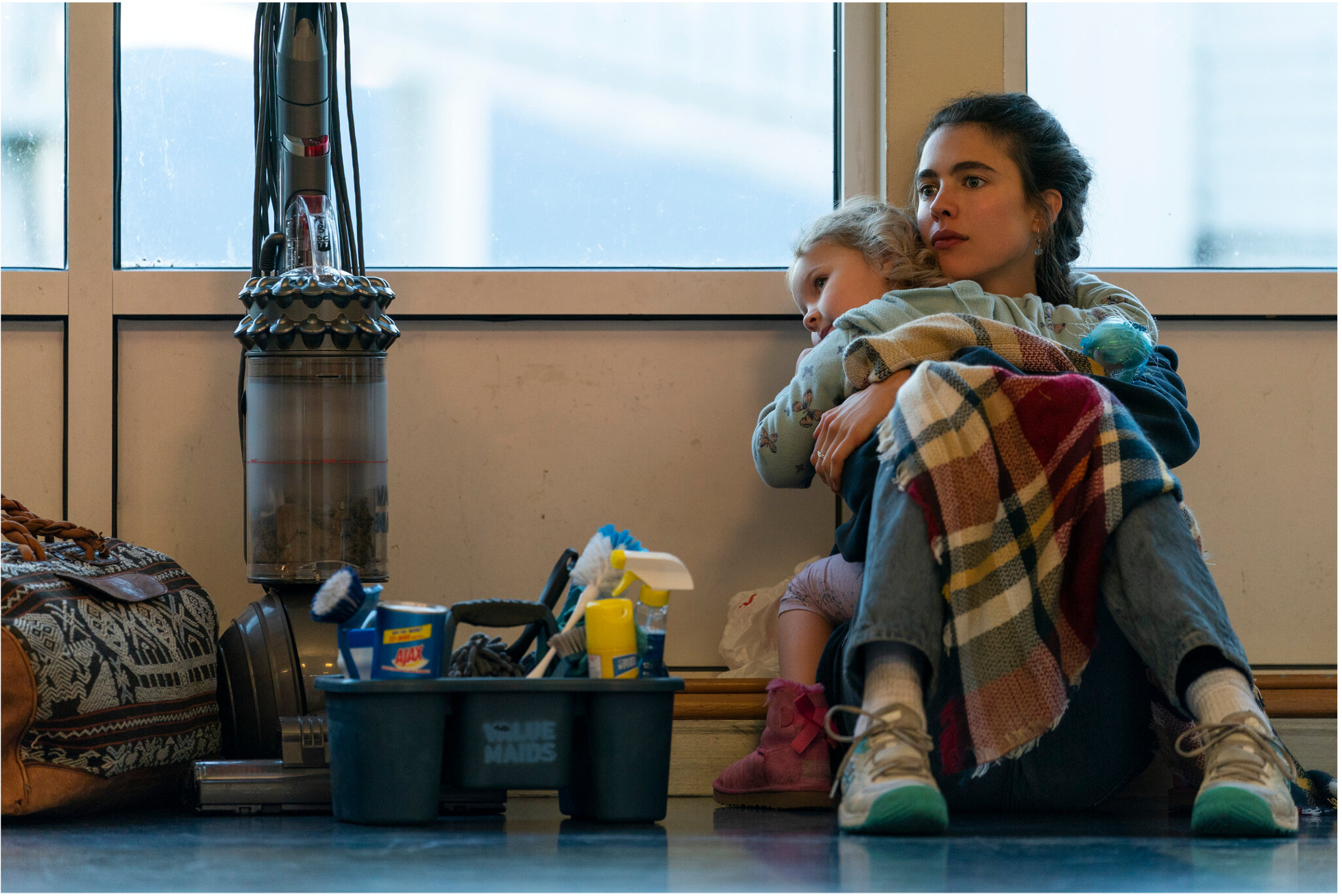 A white woman with brown hair sits on the floor holding her young child next to a box of cleaning supplies and a vacuum cleaner