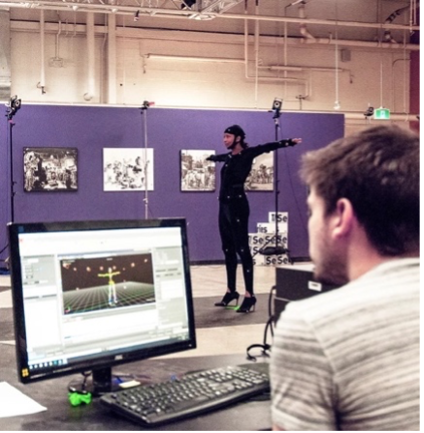 Figure 3b. A white man at a computer in front of a Black woman in a full body motion capture suit standing in a T-Pose.
