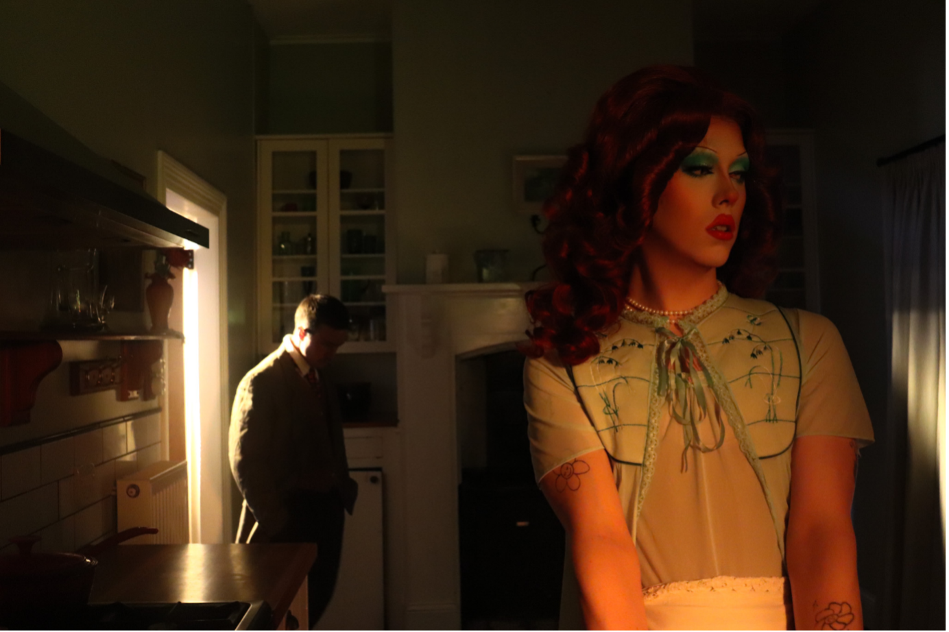 two characters standing in a dimly lit kitchen, one facing the camera forlorn and the other shadowed in the background