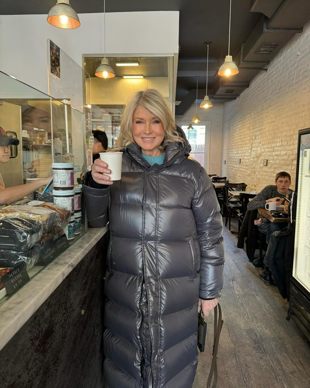 Martha Stewart holding up a cup of coffee in a coffee shop while wearing a winter jacket.