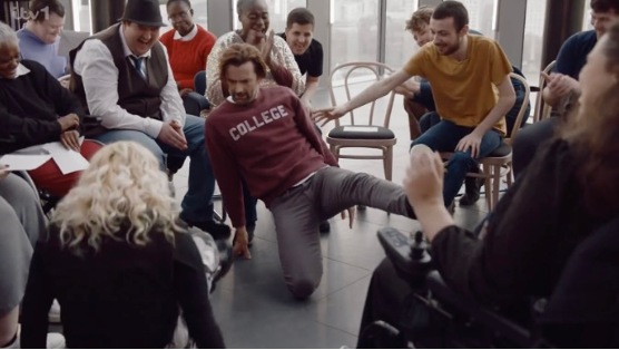 David Tennant dances in the middle of a circle of chairs while the people seated in the chairs cheer him on