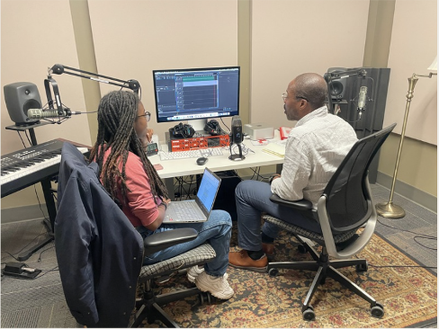 Two people sit in chairs in front of a computer monitor and recording equipment 