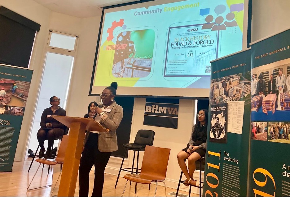 A person in a suit jacket stands in front of a podium and microphone, a slide is projected behind them with the words “community engagement” and “Black history found & forged.”