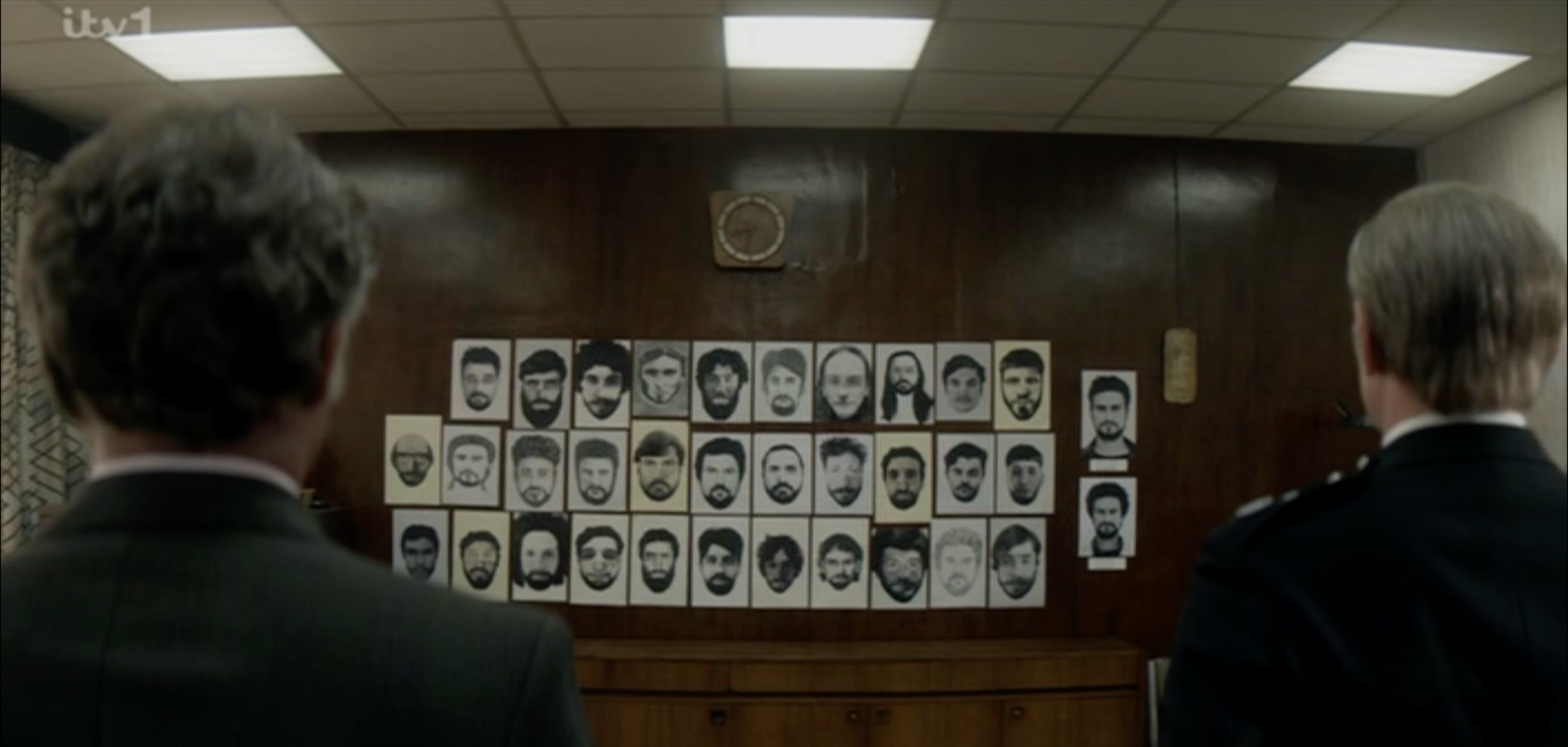 Two man stand in the foreground with their backs to the camera, staring at a wall covered in photos of different male suspects with dark hair and beards