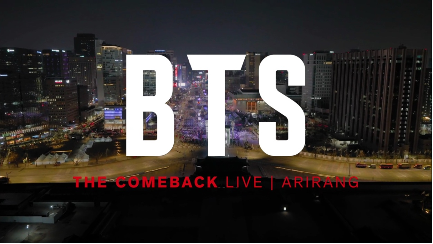 The letters B.T.S stand out in all white from the background which depicts downtown Seoul