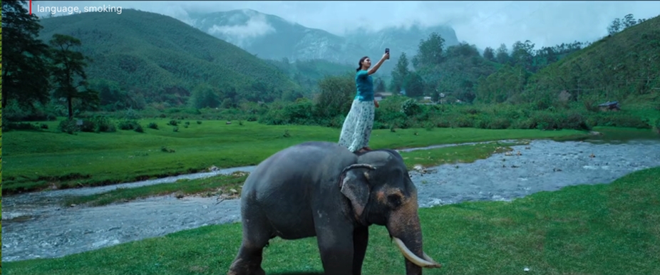 A girl standing on top of an elephant. She is raising her phone up to receive better cell reception